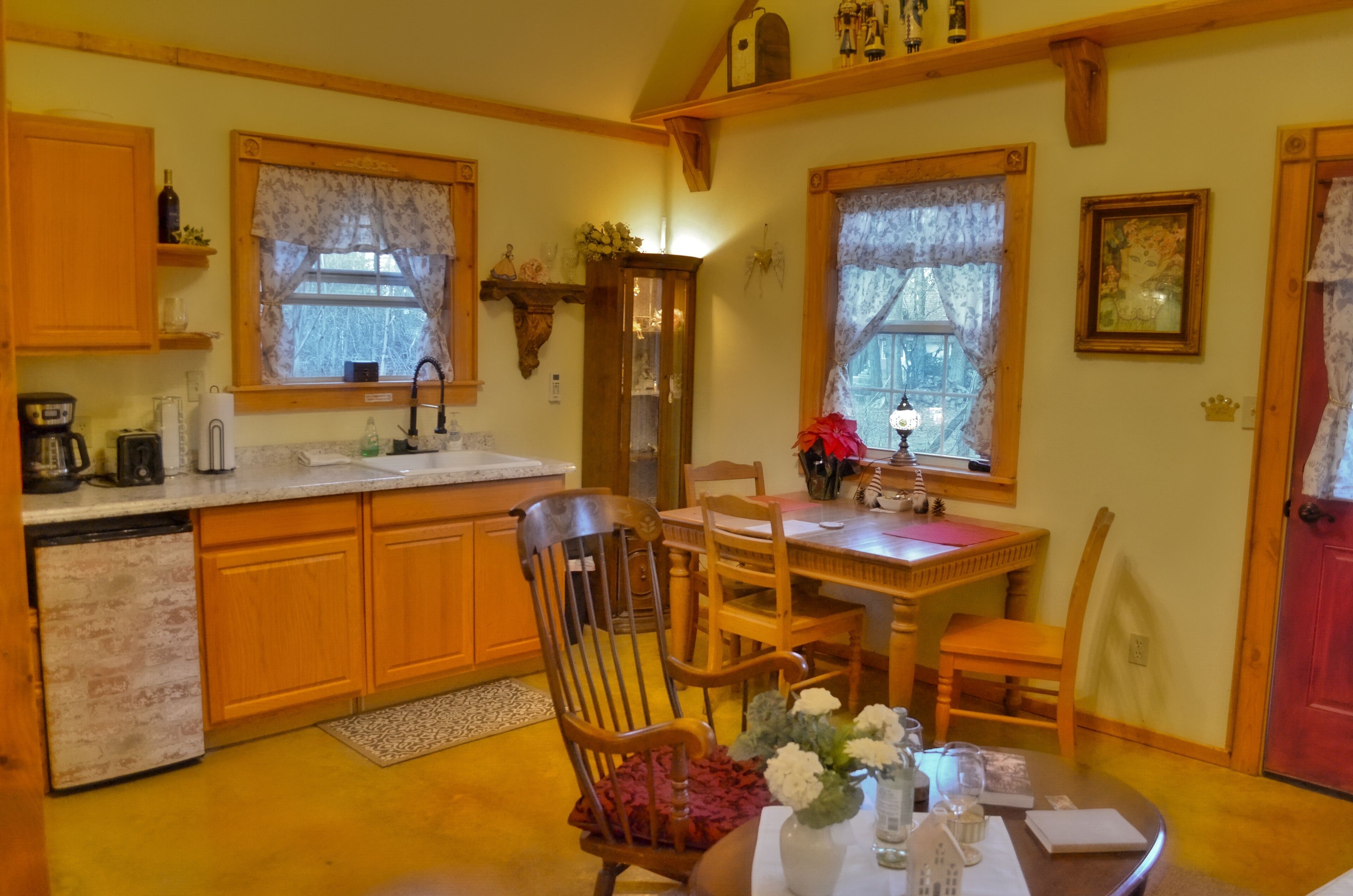 Une cuisine chaleureuse avec des meubles en bois, une table à manger entourée de chaises et une fenêtre ornée de rideaux en dentelle.