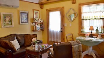 Una sala de estar acogedora con un sofá de cuero, una mesa de centro de madera y un sillón a cuadros. Se observa una estantería con libros y objetos decorativos, una ventana con cortinas y una lámpara sobre una mesita.