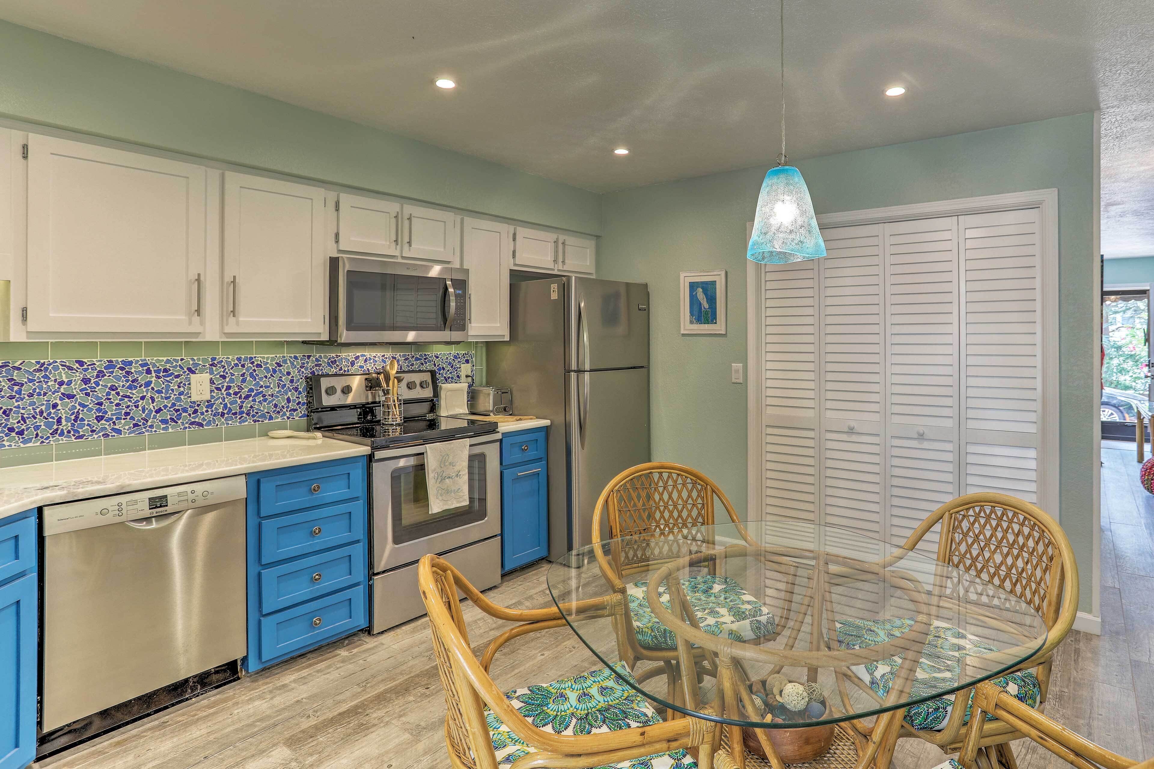 A kitchen with white cabinets, a blue countertop, a stainless steel dishwasher, a microwave, a refrigerator, and a glass dining table with wicker chairs.
