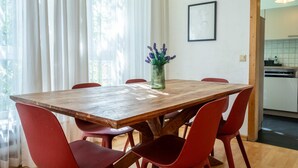 Un coin repas avec une table en bois et des chaises rouges, un vase rempli de fleurs et une cuisine visible à travers une porte ouverte.