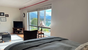 A bedroom with a large bed, a television, a chair, and a view of mountains through the window. - Studio Downtown Apt. 7 w/ Ocean & Mountain Views. (Sitka)