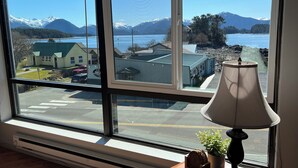 A room with a large window offering a view of a lake, mountains, and buildings. - Studio Downtown Apt. 7 w/ Ocean & Mountain Views. (Sitka)