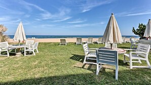 Un espace de détente aménagé sur la plage, avec des chaises longues et des tables blanches, des parasols beiges et une vue sur l’océan.