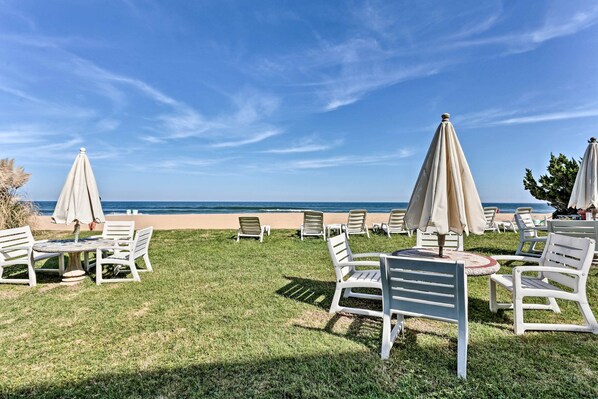 A beachside lounge area with white lounge chairs and tables, beige umbrellas, and a view of the ocean. - Virginia Beach Studio w/ Balcony + Pool View! (Virginia Beach)