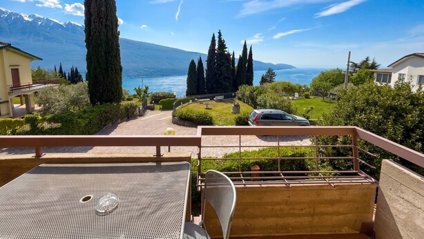 A balcony with a metal table and chair, overlooking a scenic view of a lake, mountains, and a car parked in the distance.