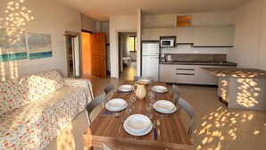 A dining area with a wooden table set for four, a sofa with a floral cover, and a kitchen in the background.