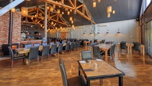 A spacious dining area with high ceilings, exposed wooden beams, and a brick wall. Tables are set with chairs, and a kitchen area is visible in the background. - Dubbo Rhino Lodge (Dubbo)
