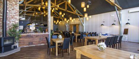 A spacious restaurant with high ceilings, exposed beams, and numerous hanging pendant lights. Tables and chairs are arranged for dining.