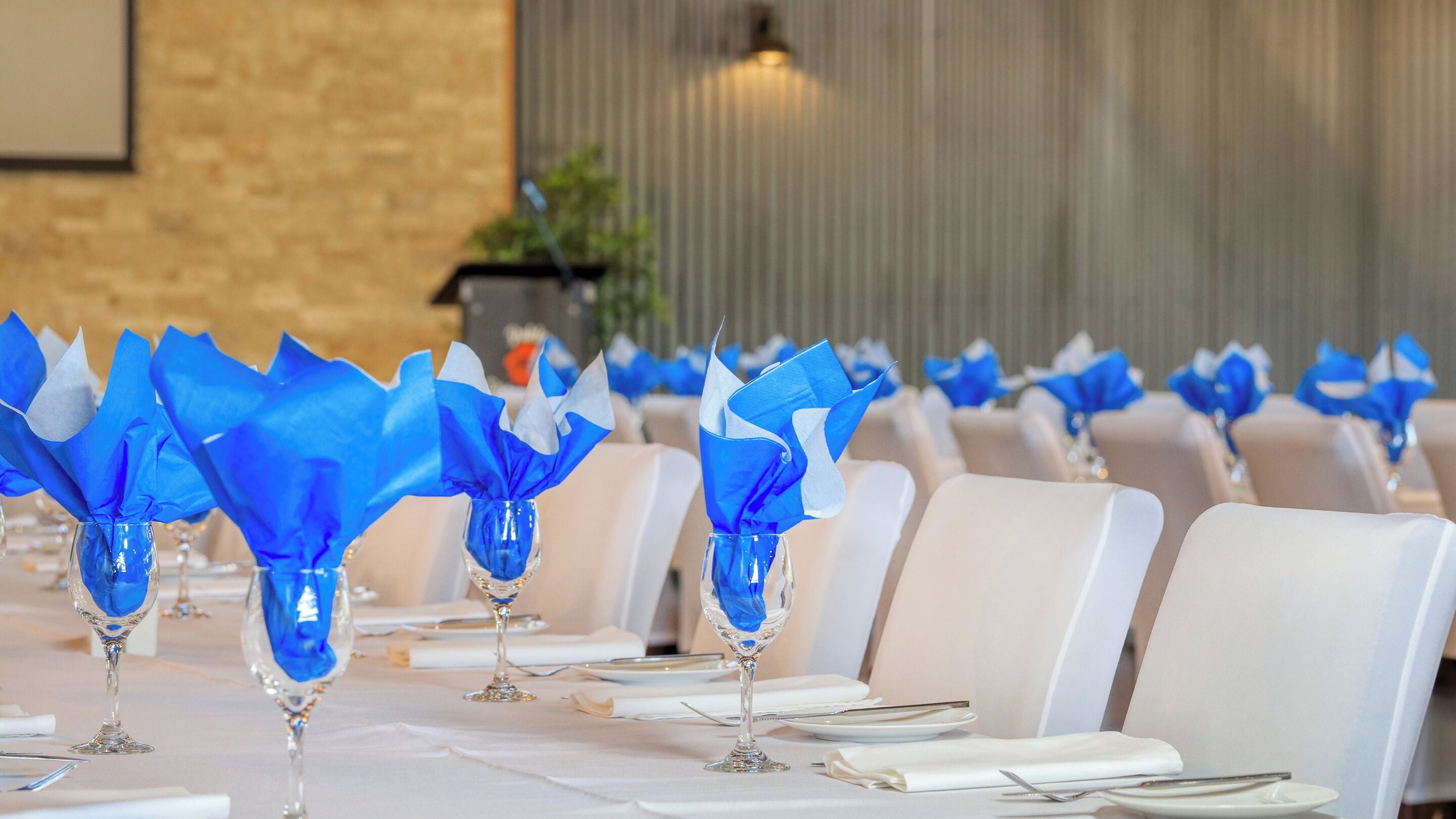 A row of tables with blue napkins and clear glass vases, set against a brick wall and a vertical striped pattern.