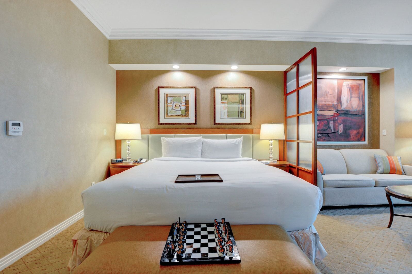 A hotel room with a large bed, two bedside tables with lamps, a sofa, a chessboard, and two framed pictures on the wall.