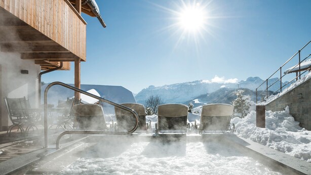 Un jacuzzi con vapor, rodeado de nieve, con un edificio de madera y montañas al fondo.
