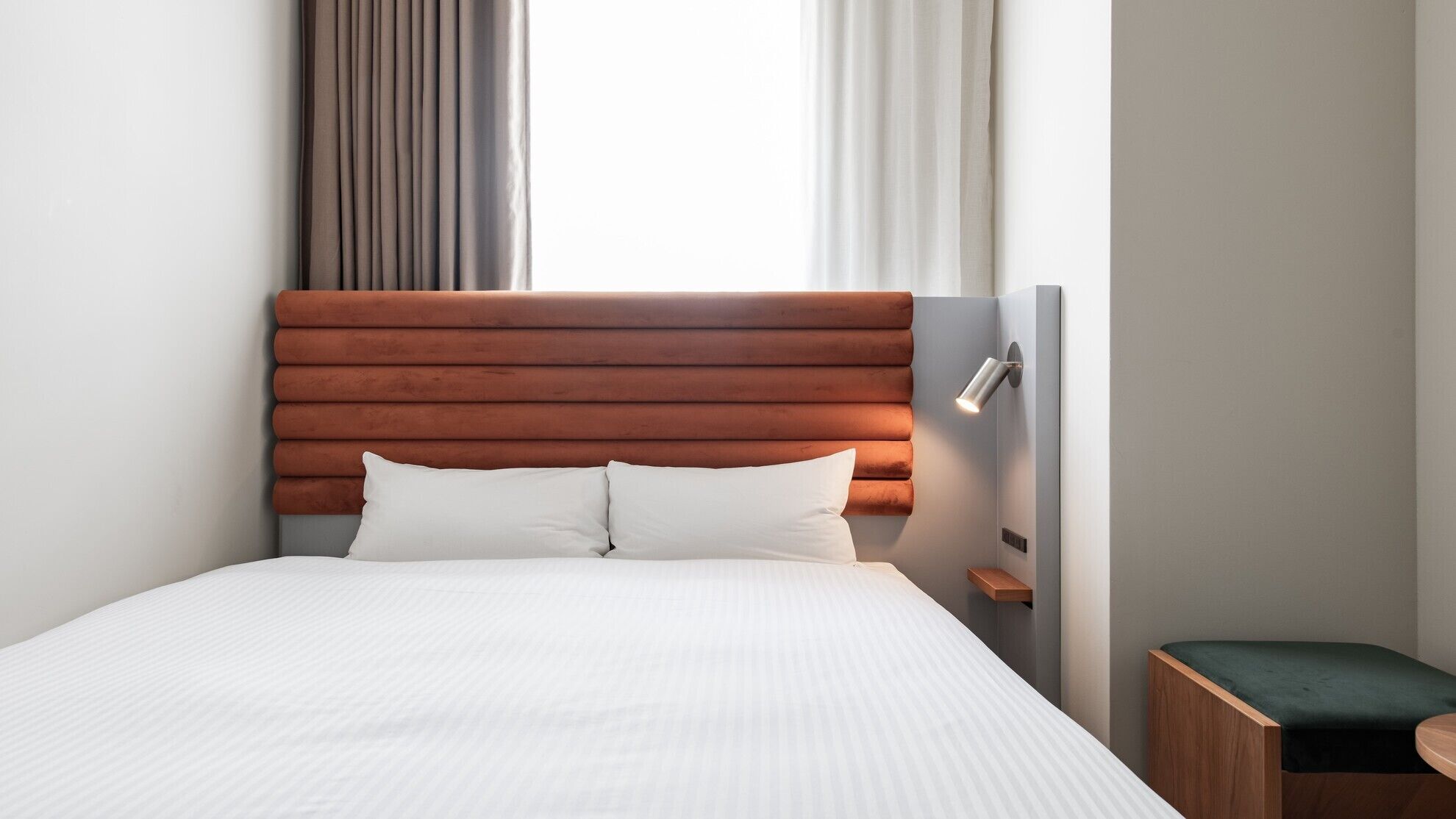 A hotel room with a large bed, a wooden headboard, a bedside table, a green bench, and a wall-mounted lamp.
