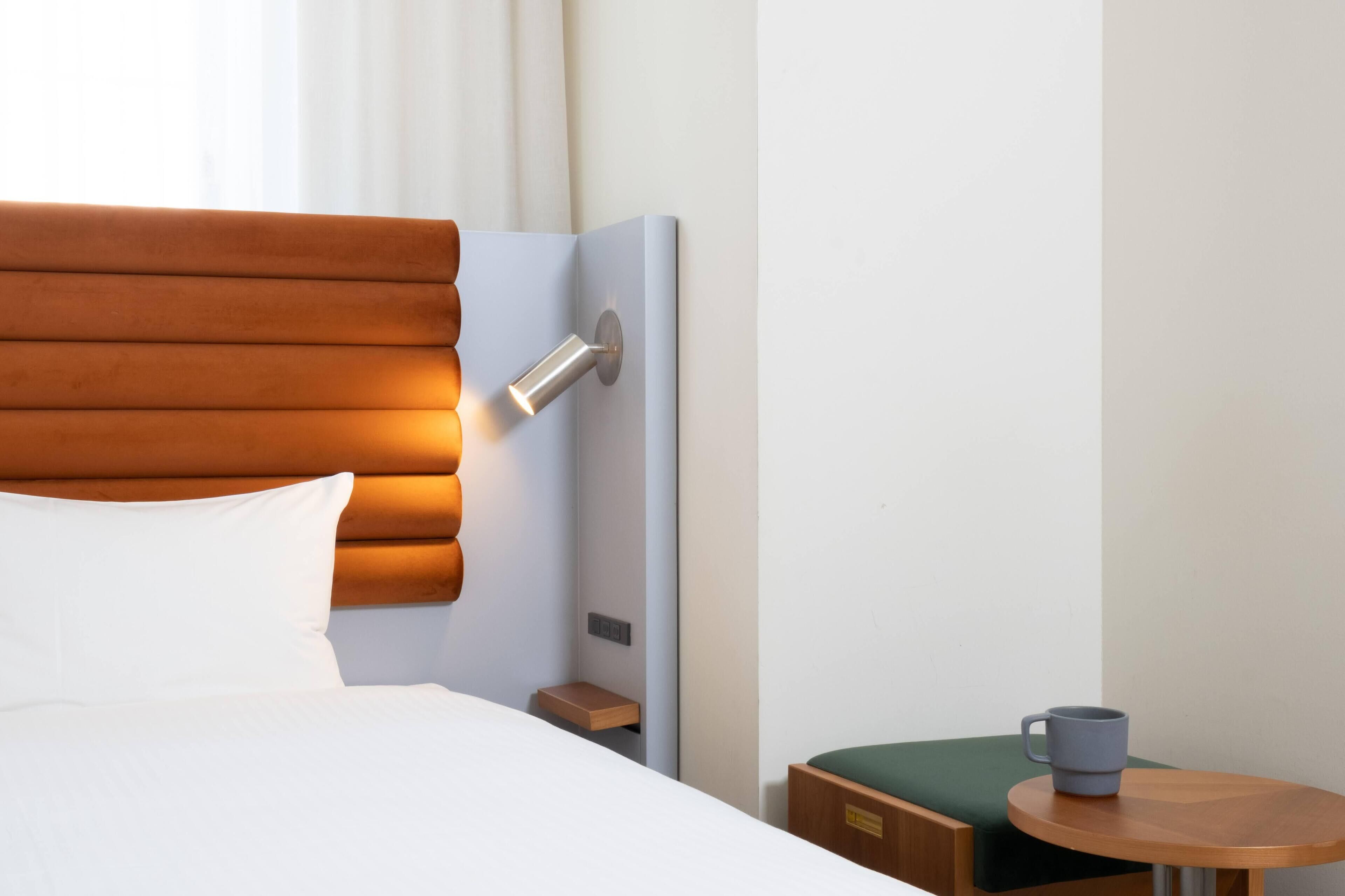 Una cama bien hecha con cabecera, una mesita de noche con una taza, y una pequeña mesa con un cojín verde.