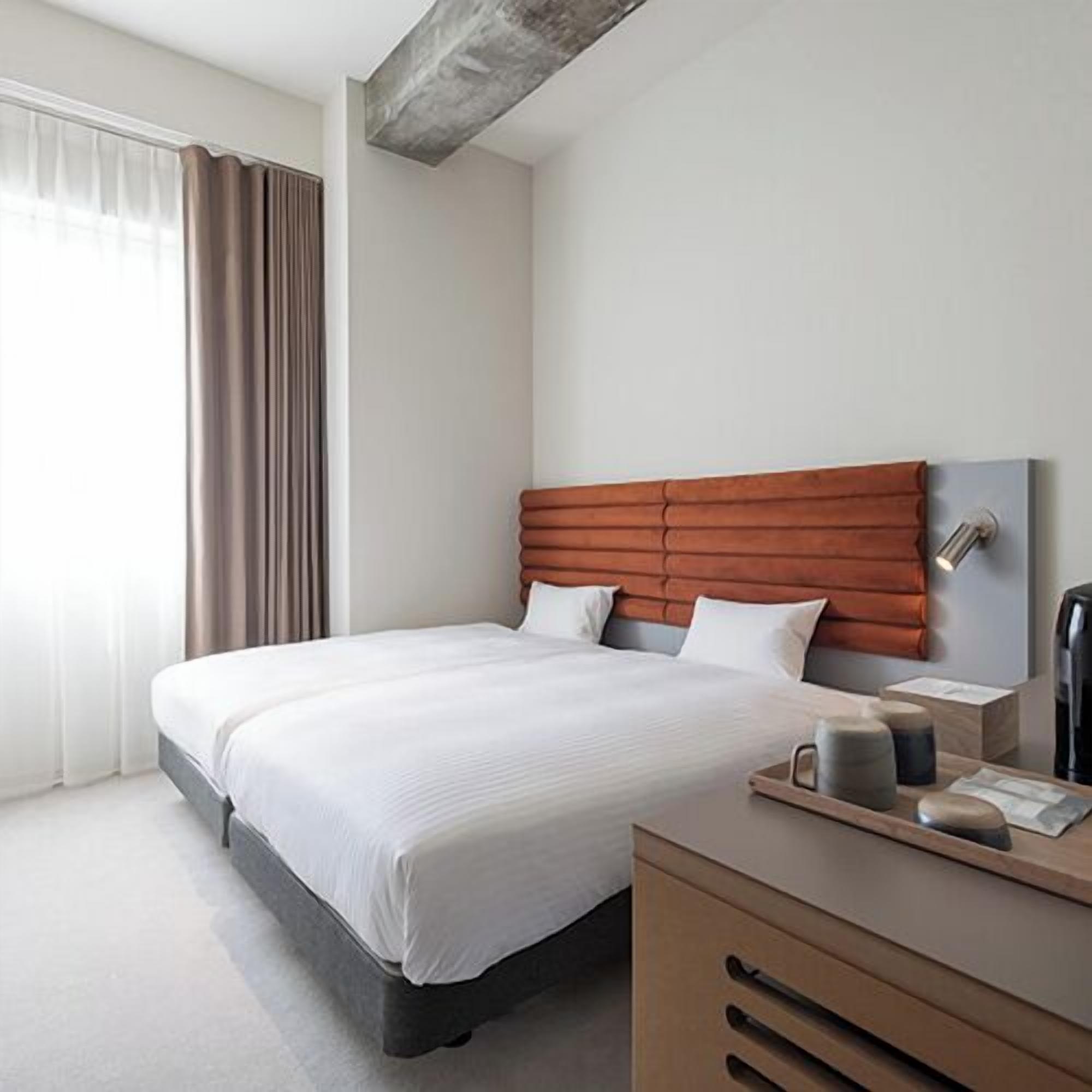 Ein modernes Hotelzimmer mit einem großen Bett, einem hölzernen Kopfteil, einem Nachttisch mit Tablett und einem Fenster mit Vorhängen.