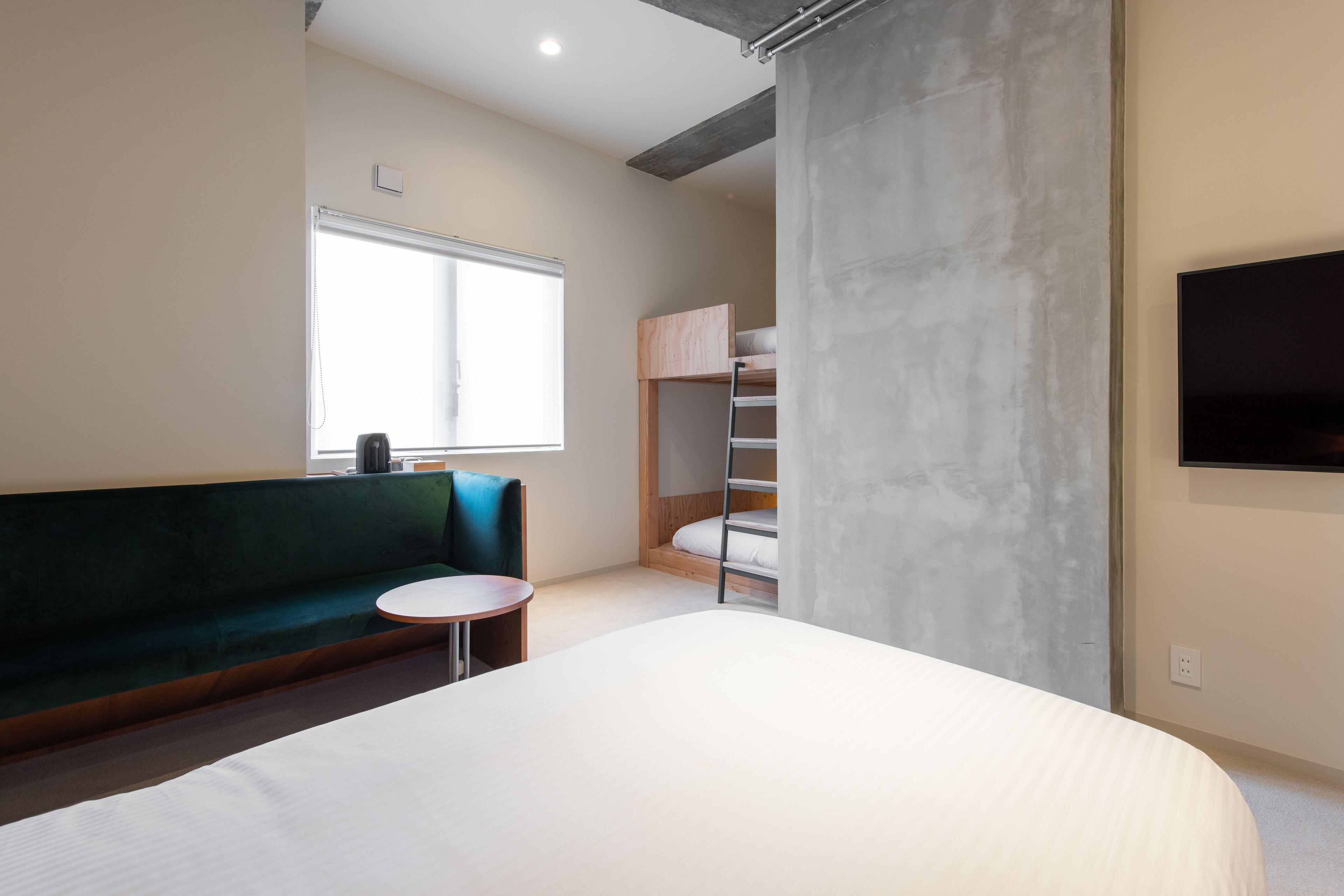 A modern hotel room with a large bed, a green sofa, a small round table, a bunk bed, and a TV mounted on the wall.