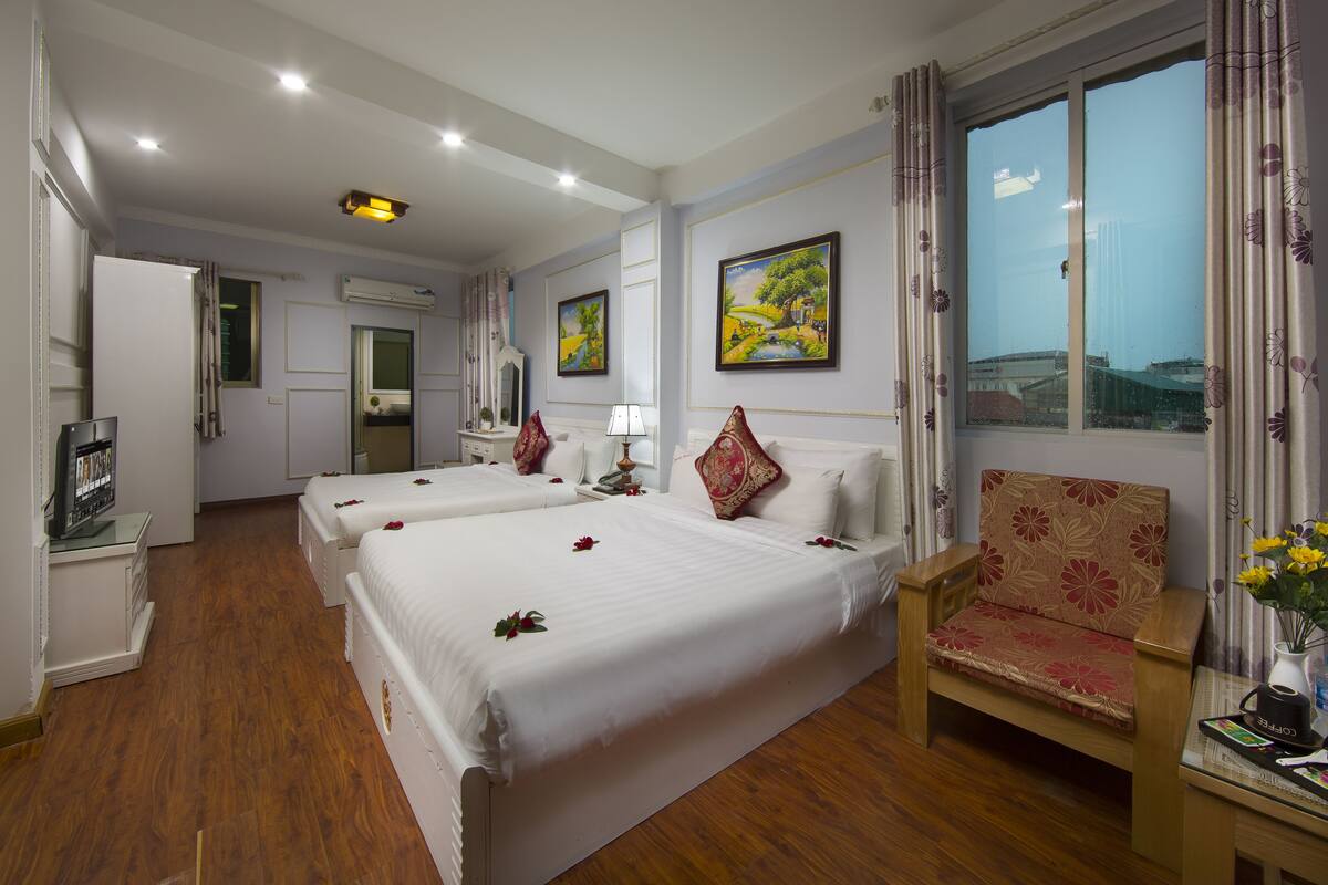 A hotel room with two beds, a wooden chair, a desk with a lamp, and a window with a view.