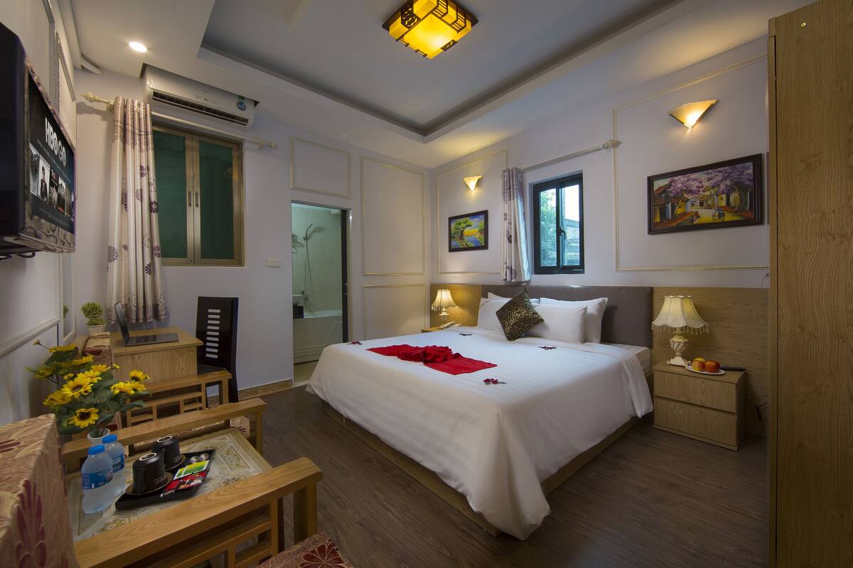 A hotel room with a large bed, bedside tables, a desk with a chair, a window with curtains, and wall-mounted artwork.