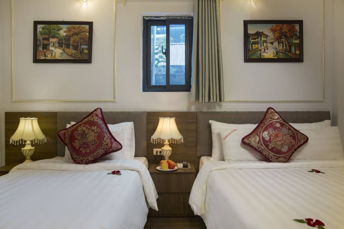 A hotel room with two beds, each with a red decorative pillow, a bedside table with a lamp, and a small plate of fruit. There are two framed paintings on the walls, and a window with curtains.