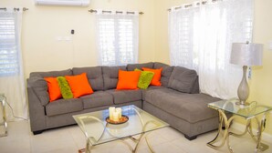 A living room with a grey sectional sofa, orange and green cushions, a glass coffee table with candles, and a lamp on a glass side table. - Ocean View Villa at Coolshade Ocho Rios (St. Ann's Bay)