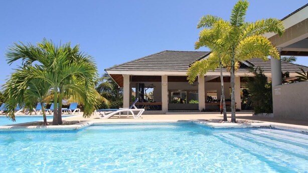A swimming pool with lounge chairs and palm trees, surrounded by a building with a covered patio area. - Ocean View Villa at Coolshade Ocho Rios (St. Ann's Bay)