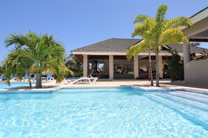 Une piscine entourée de chaises longues et de palmiers, le tout bordé par un bâtiment doté d’un patio couvert.
