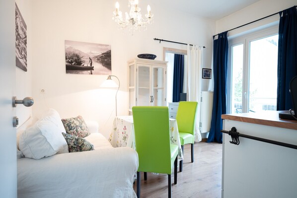 Une pièce comprenant un lit, une table à manger avec des chaises, un buffet, un lustre et une fenêtre avec des rideaux.