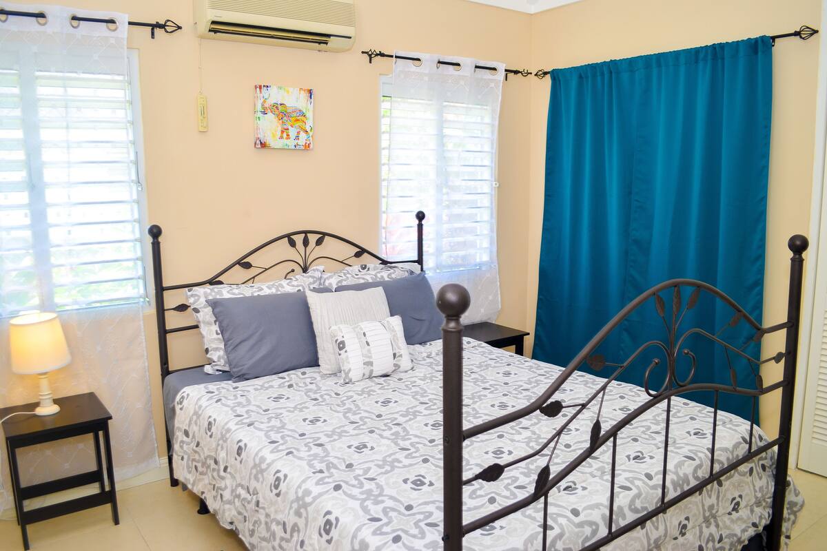 A neatly arranged bedroom with a bed, a bedside table with a lamp, a window with a curtain, and an air conditioning unit on the wall.