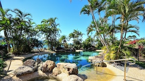 Una piscina cristalina rodeada de rocas y palmeras, con una barandilla metálica en primer plano.
