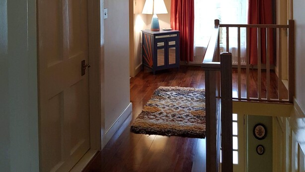 A hallway with wooden flooring, a door on the left, a lamp on a side table, and a staircase with a railing on the right. - Comfortable Home in a beautiful part of Dingle Town (Dingle)