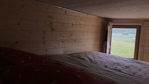 Un dormitorio con techo y paredes de madera, una cama individual con colcha roja y blanca, y una ventana que ofrece vistas a un campo verde.