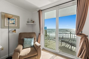 A room with a view of the ocean, a chair with a cushion, a floor lamp, and a framed picture on the wall.