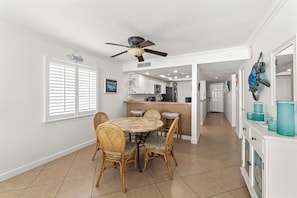 A dining area with a round table and four chairs, a ceiling fan, a kitchen in the background, and a hallway leading to a door.