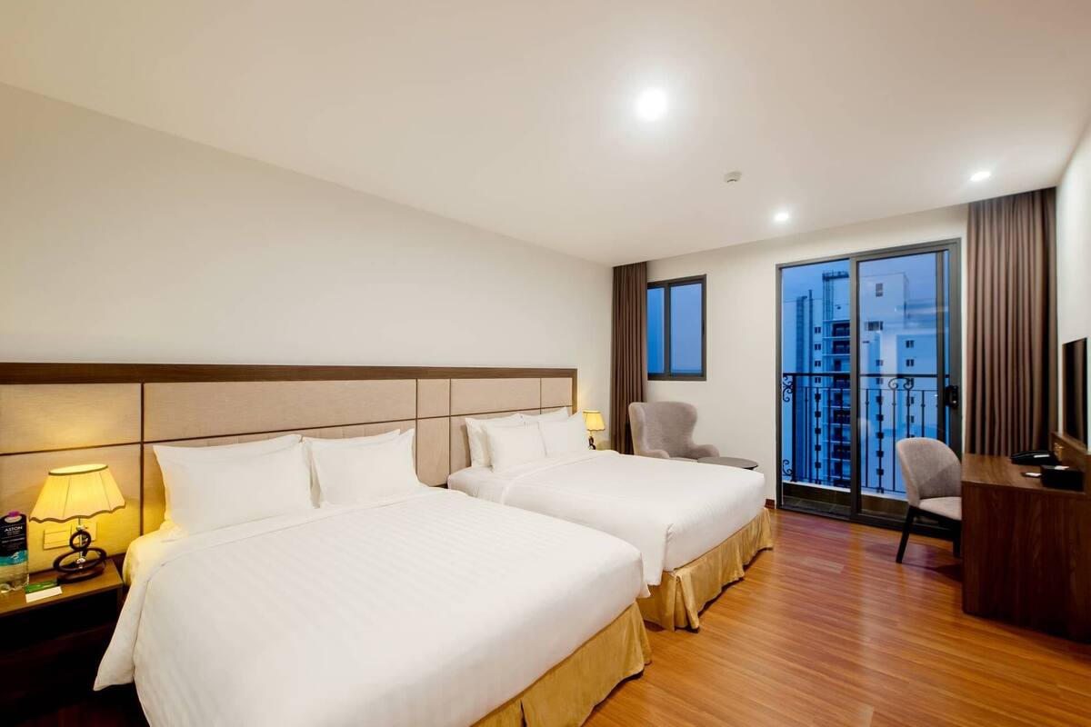 A hotel room with two beds, a desk, a chair, and a view of the cityscape through a large window.