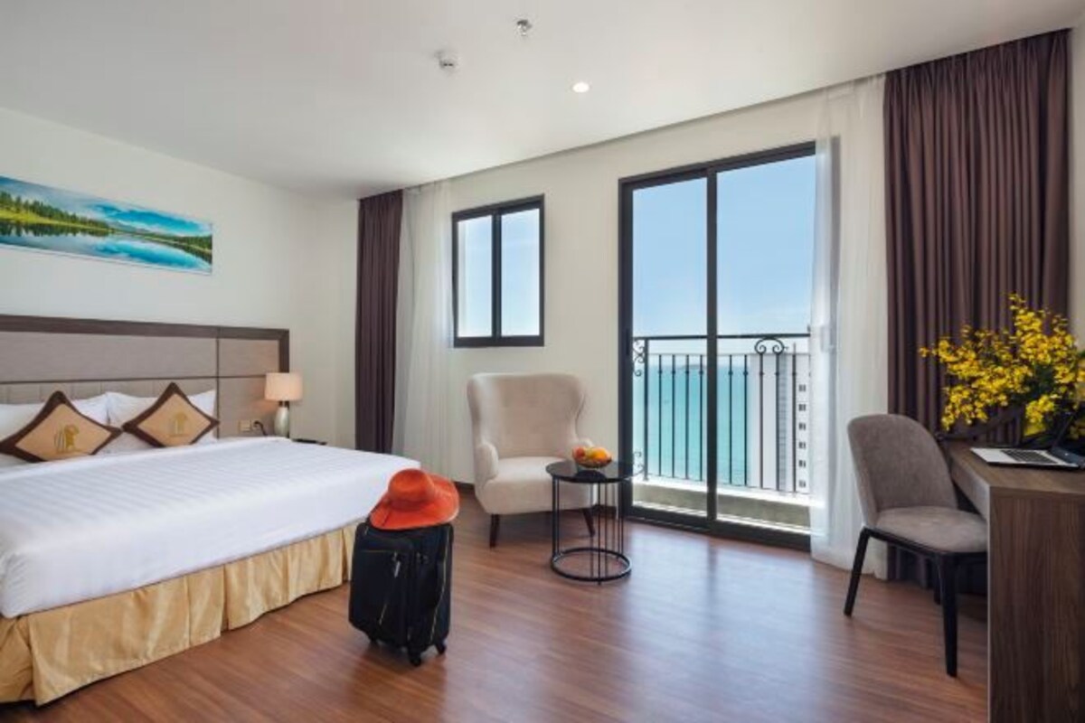 A hotel room with a bed, a desk, a chair, a small table with a lamp, and a view of the sea through large windows.