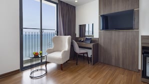 A modern hotel room with a large window, a desk, a chair, a flat-screen TV, and a small table with fruit. - Daphovina Hotel (Bac Nha Trang)
