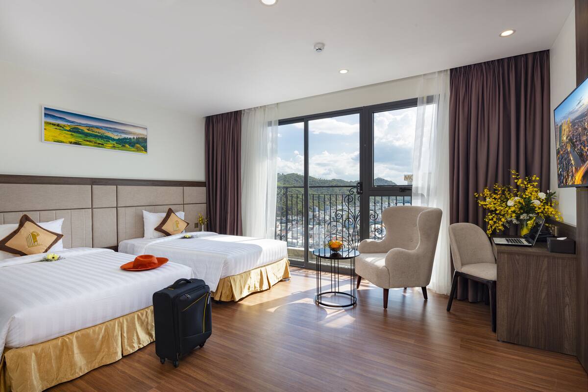 A hotel room with two beds, a desk, a chair, a suitcase, and a balcony with a city view.
