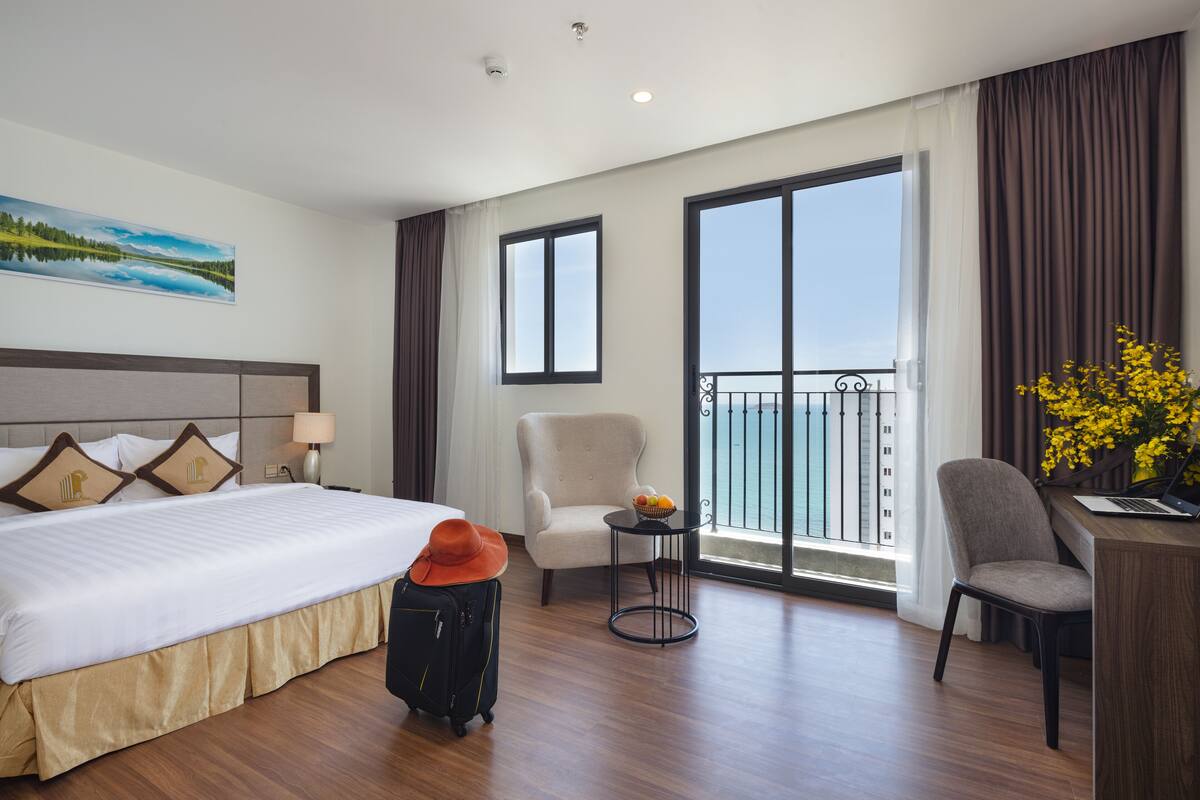 A hotel room with a large bed, a desk, a chair, a small table with a fruit bowl, and a balcony with a view.