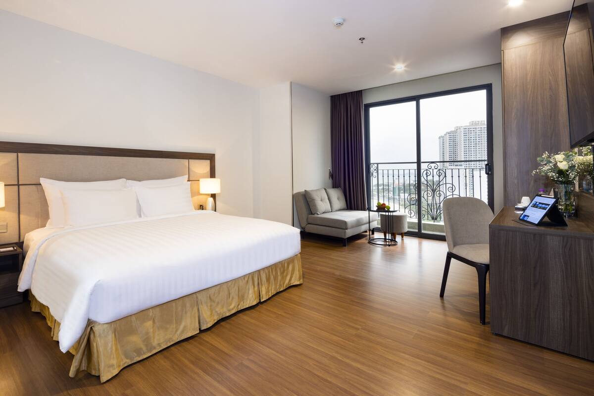 A hotel room with a large bed, a desk, a sofa, and a balcony with a view of buildings.