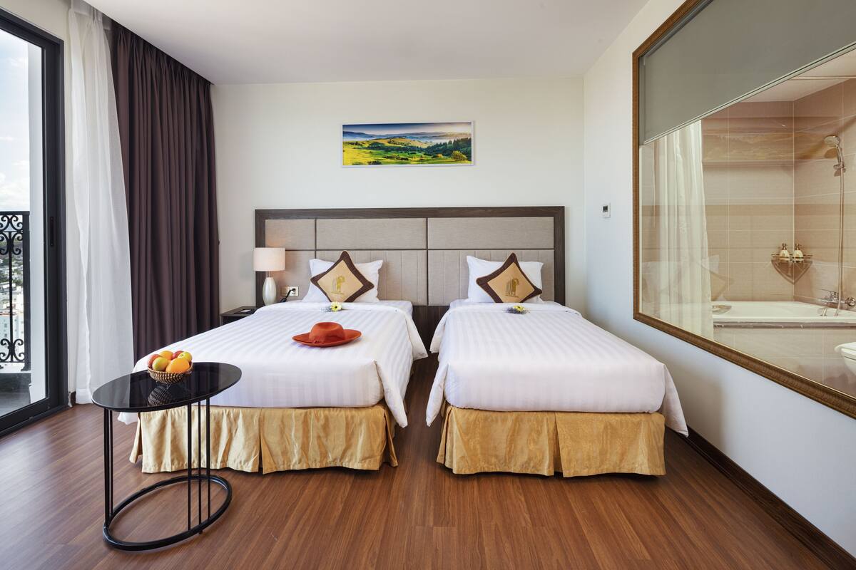 A hotel room with two beds, a small table with fruit, a painting on the wall, and a large mirror.