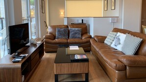 A living room with a brown leather sofa, a glass coffee table, a TV on a wooden stand, and a window with a view of the outside. - Diamond Dust Furano (Furano)