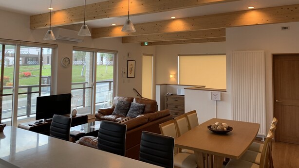 A modern living room with a wooden ceiling, a leather sofa, a dining table, and a large window offering a view of the outdoors. - Diamond Dust Furano (Furano)