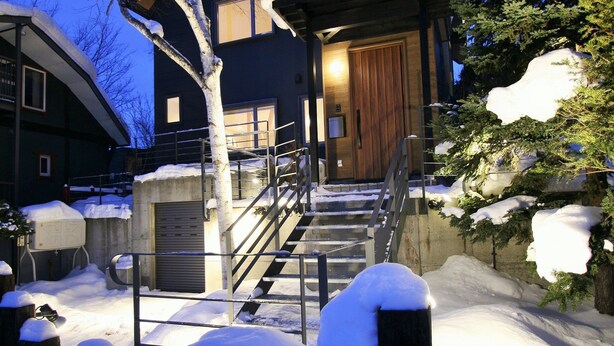 A modern house with a wooden door, a staircase leading to a garage, and snow-covered surroundings. - Asanoha (Furano)