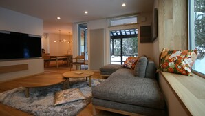 A modern living room with a sofa, a TV, a dining area, and large windows. - Asanoha (Furano)