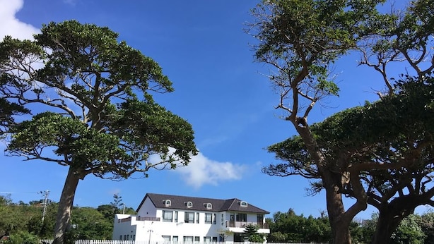 A two-story white building with a dark roof, surrounded by a white picket fence, and large trees in the foreground. - Summer Homestay (Hengchun)