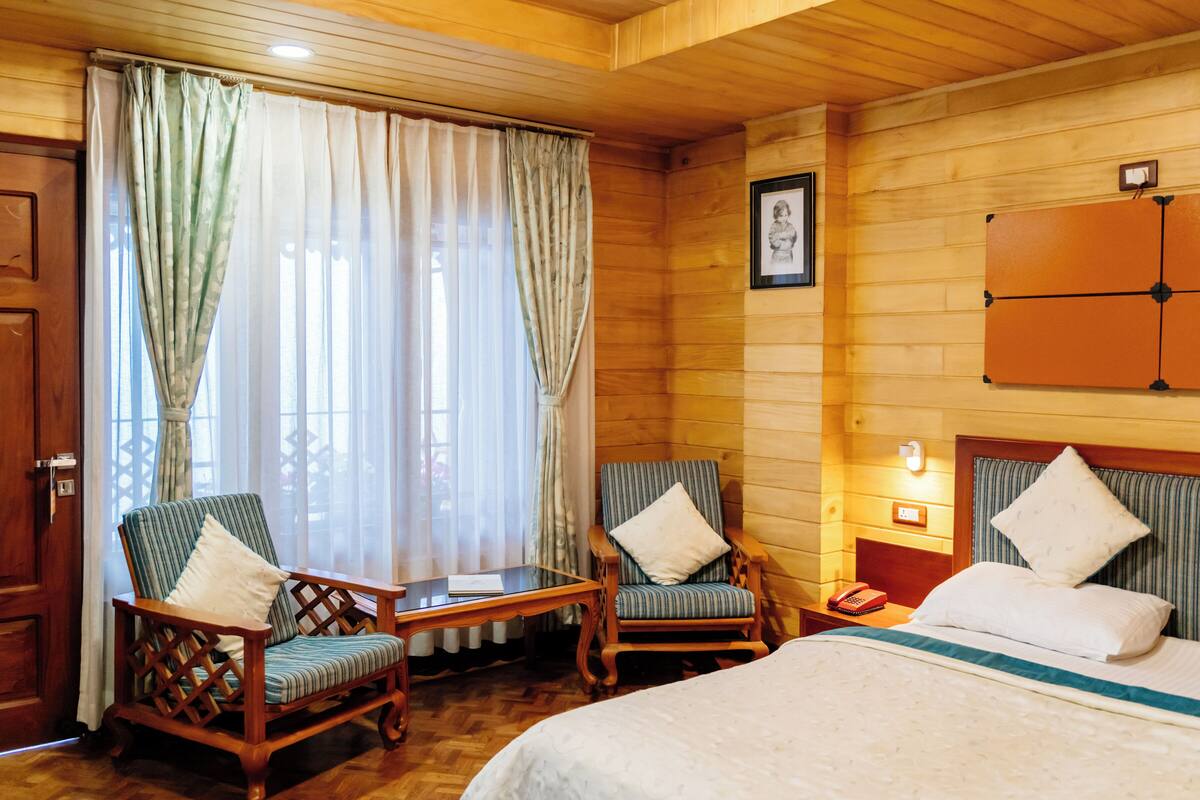 Een houten kamer met twee houten stoelen, een bed, een nachtkastje en een lijst met een afbeelding aan de muur.