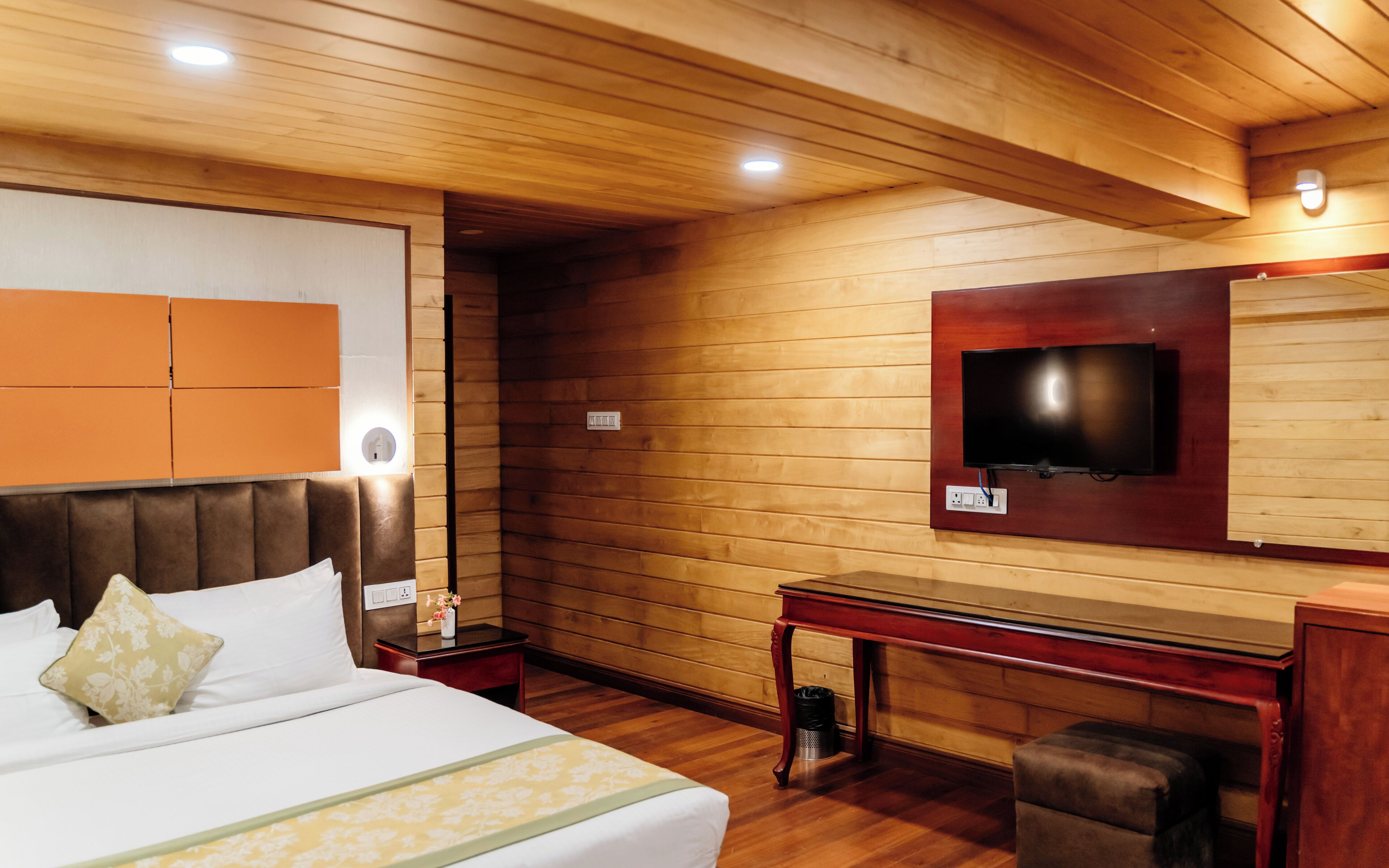 A hotel room with a wooden ceiling, a bed with white bedding, a bedside table, a TV mounted on the wall, and a small desk with a chair.