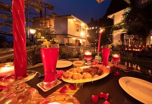 Een buitenterras met een gedekte tafel voor twee personen, met een bord eten en twee glazen rode drank, omringd door kaarsen en rozenblaadjes.