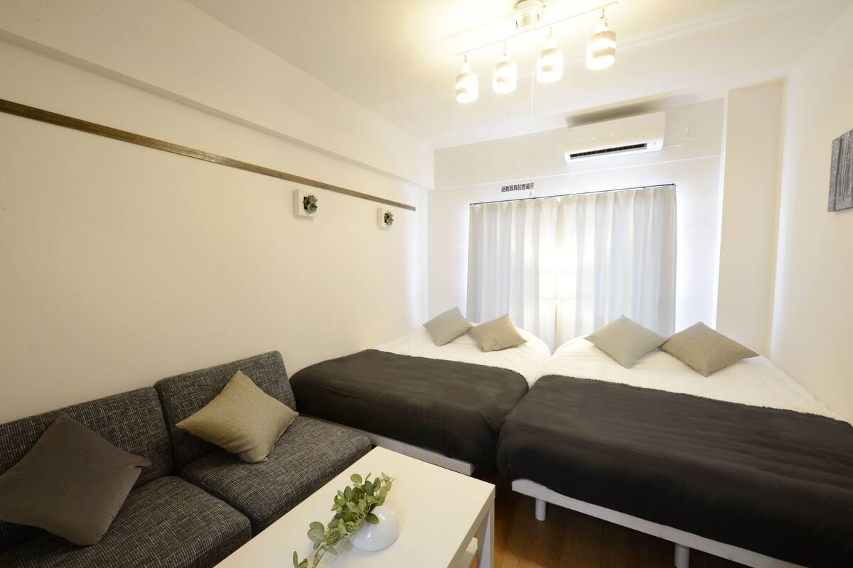 A room with a sofa, two single beds, a small table with a plant, and a window with curtains.