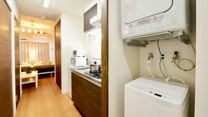 Una cocina compacta con microondas, estufa y calentador de agua. Se observa un pequeño comedor con una mesa y sillas al fondo.
