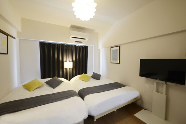 A hotel room with two beds, a flat-screen TV, an air conditioning unit, and a window with curtains.